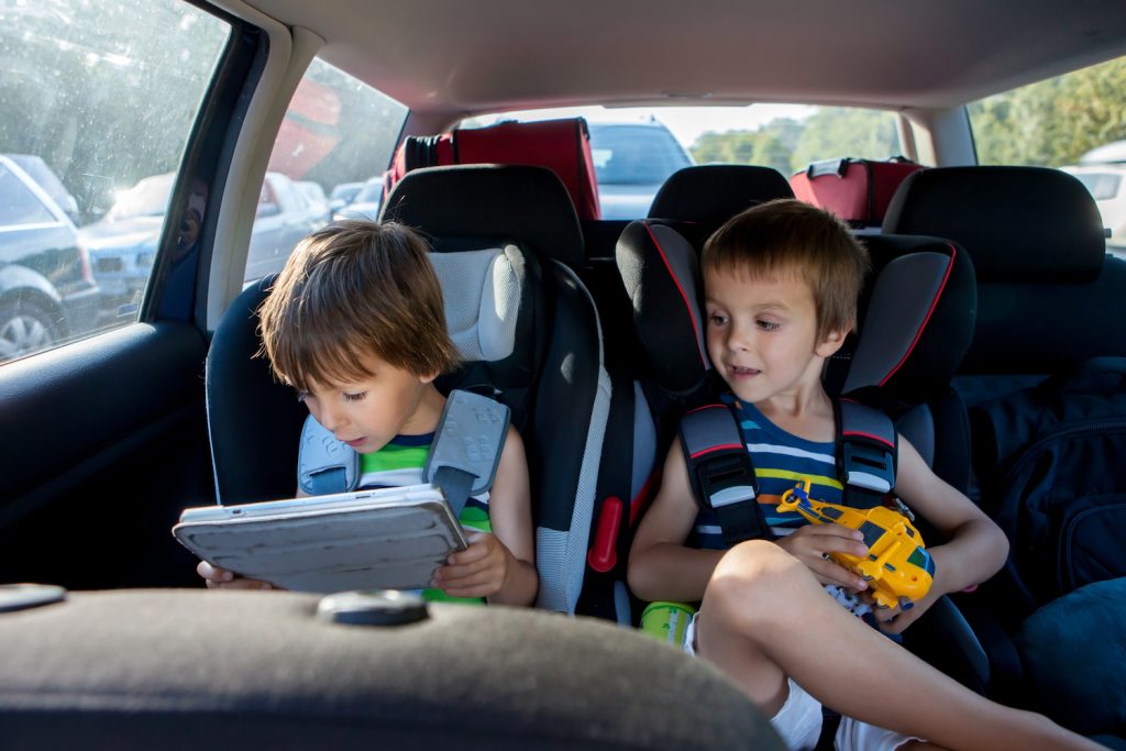 Two Boys In Children Car Seats, Traveling By Car And Playing With Toys And Tablet