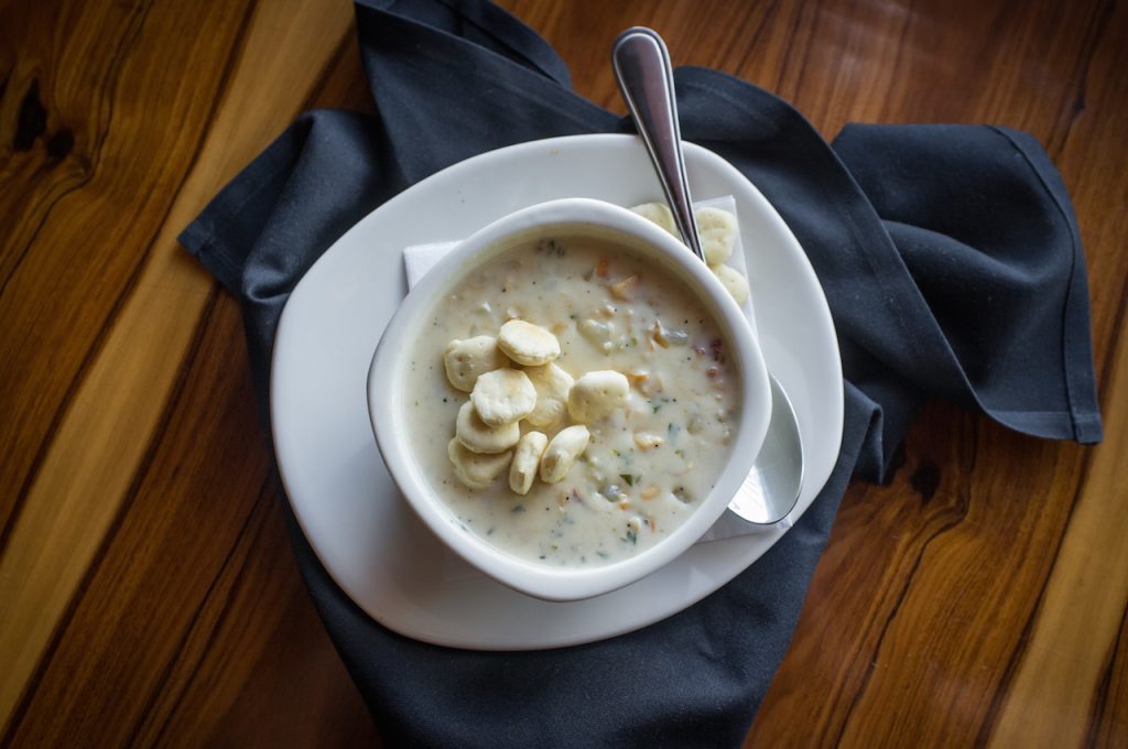 a cup of new england clam chowder, topped with crackers, on a plate with a spoon and blue napkin 