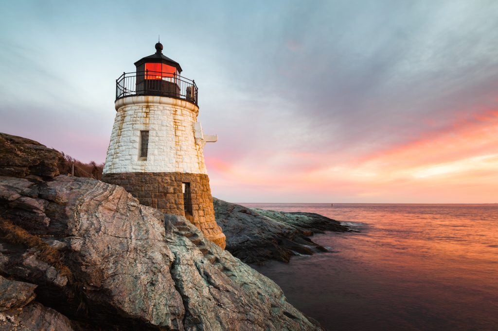 Small Castle Hill Lighthouse sits on the rocky coastline of Newport, Rhode Island at sunset with the waves slowly rushing across the rocks.